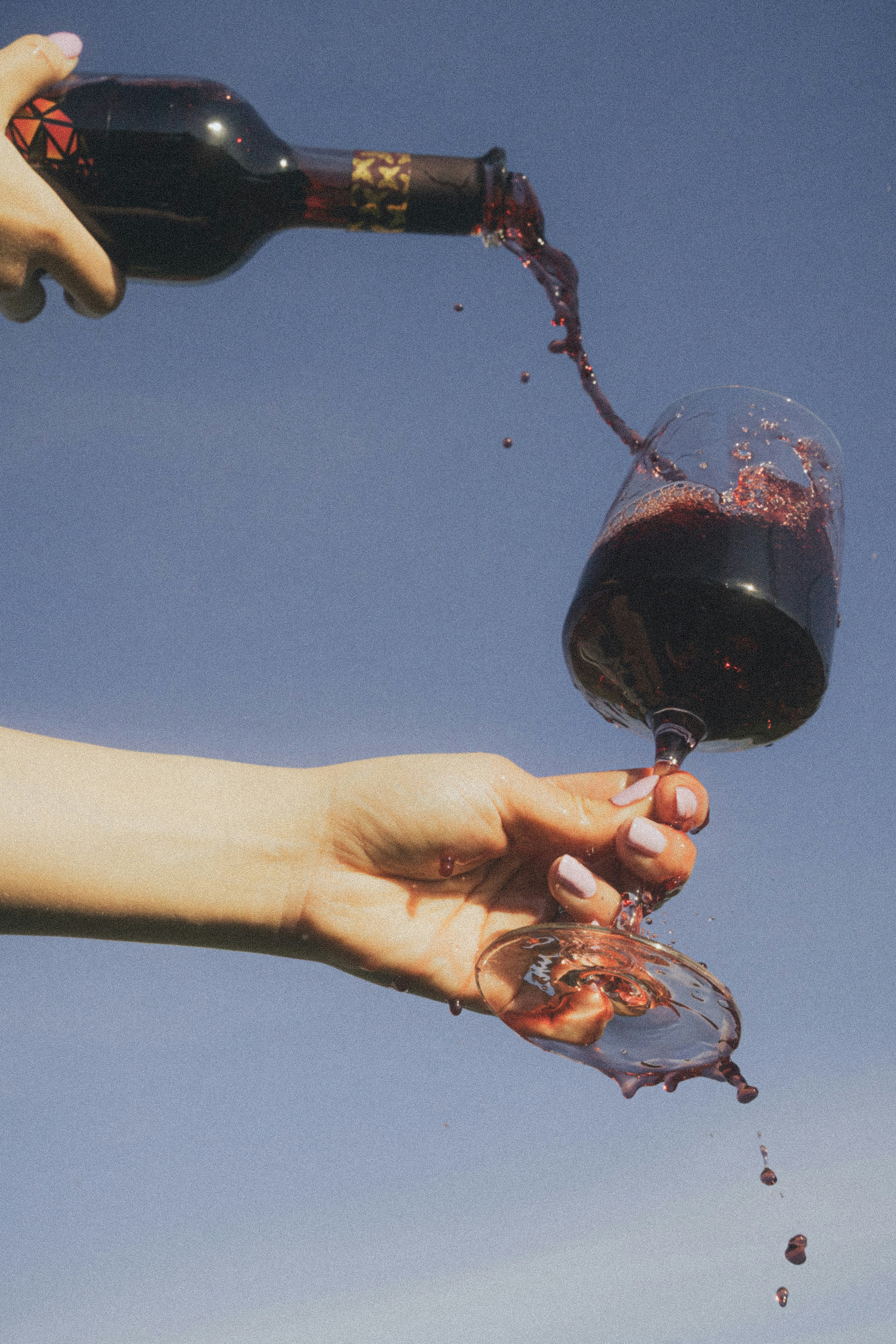 A hand holding a tipping wine glass with red wine spilling, against a blue sky