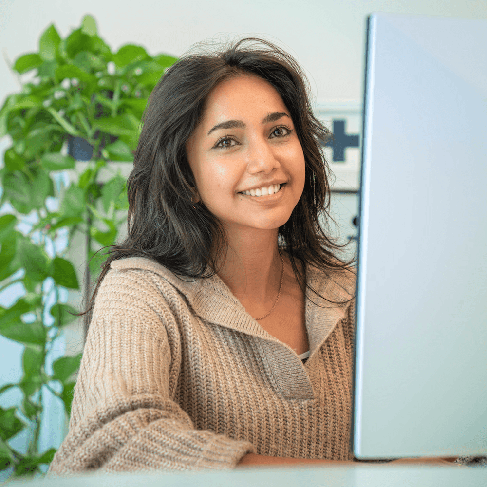 Frau mit braunem, schulterlangem Haar und beigem Strickpullover sitzt vor einem silbernen Laptop, im Hintergrund grüne Pflanze und weiße Wand mit schwarzem Kreuzsymbol