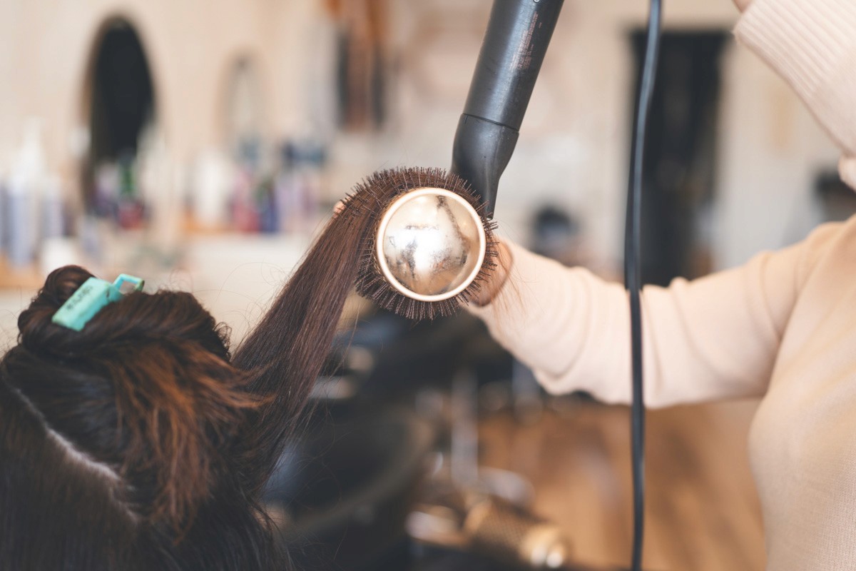 Close-up of a hairstylist blow drying dark hair with a round brush and dryer, creating volume and a smooth, polished finish in the salon.