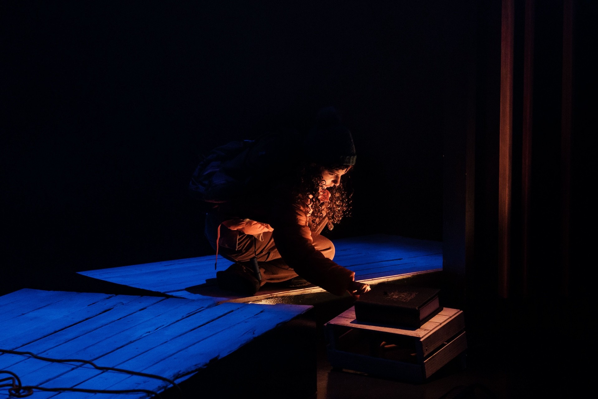 A performer in an orange puffer jacket crouches beside a wooden platform in a darkened space, illuminated by a small warm light as they examine an object inside a crate.