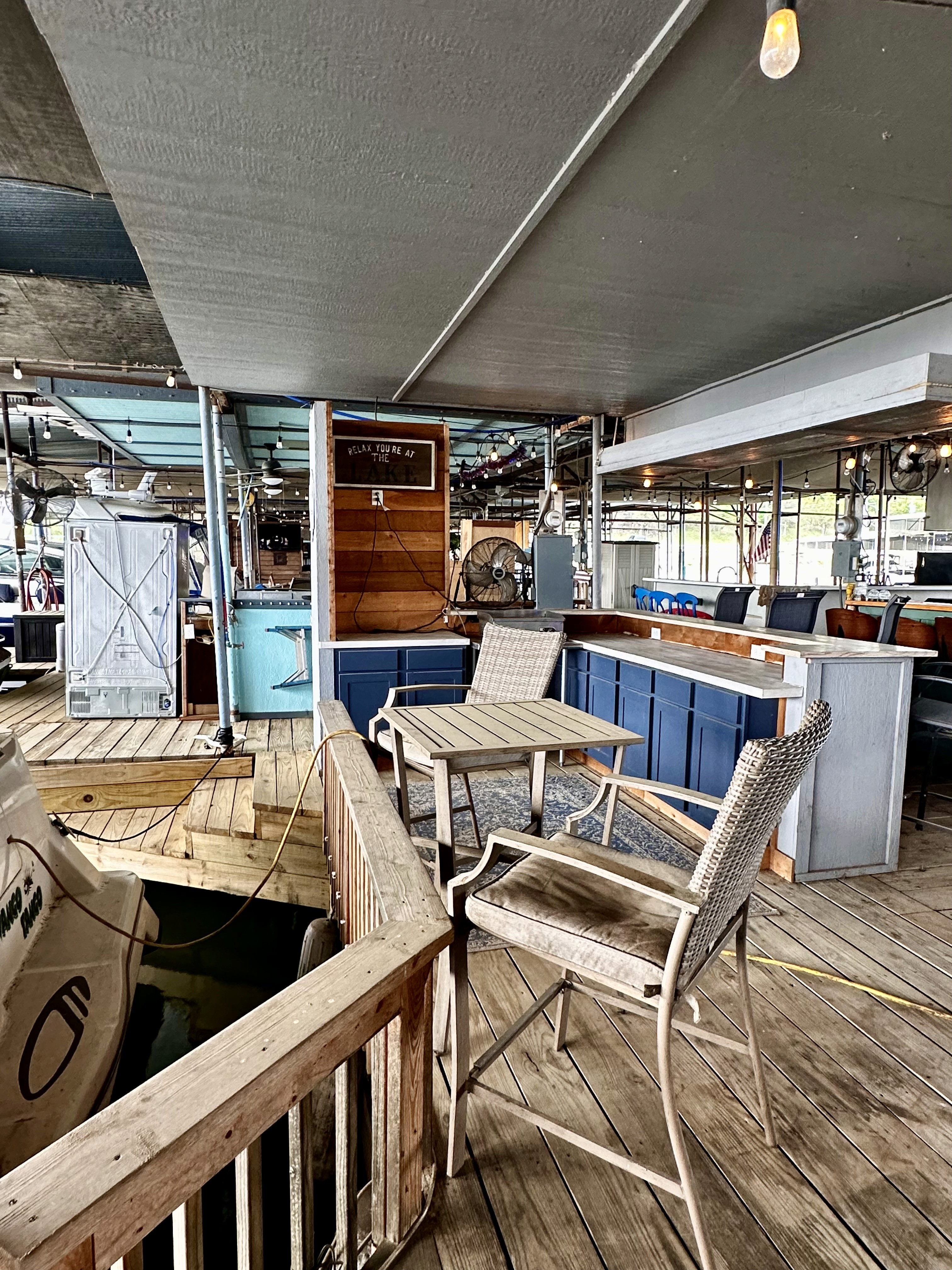 An outdoor marina lounge area features a wooden deck with woven chairs and a small table, adjacent to a counter set against a backdrop of berthed boats and serene waterfront views.