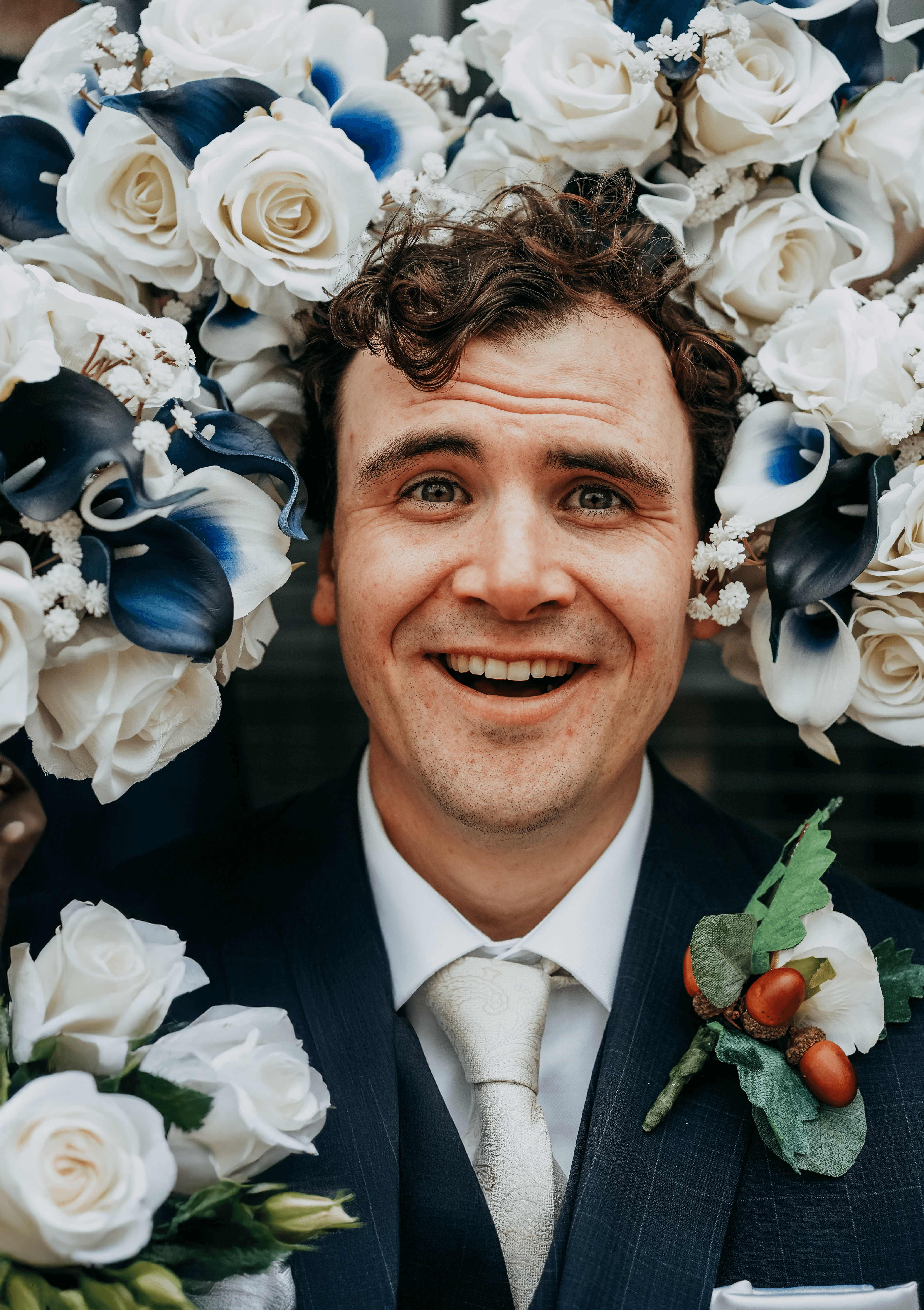 A fun, close-up portrait of the groom smiling with excitement at the camera. His head is framed by bouquets of white roses and blue calla lilies, creating a floral border around him.