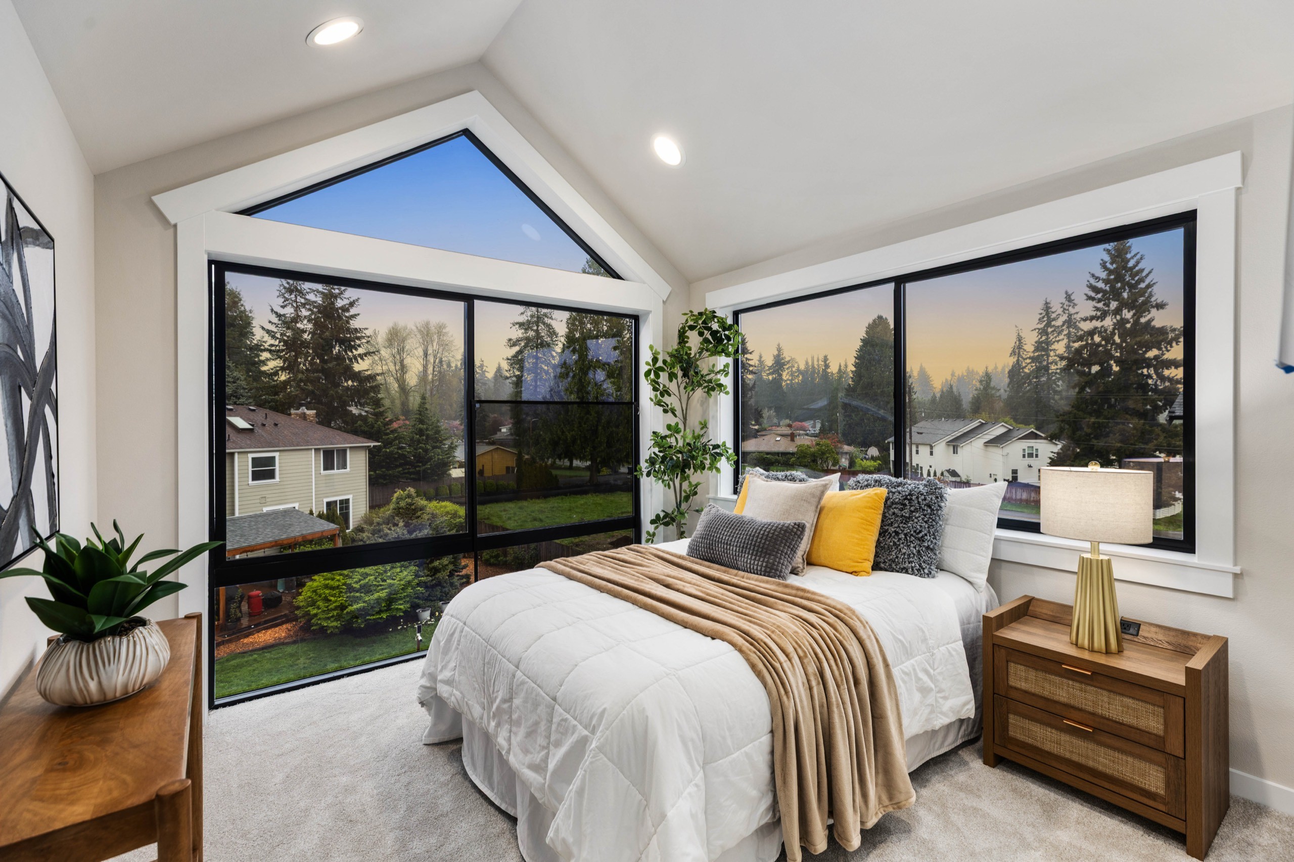 Stunning luxury primary bedroom with dramatic floor-to-ceiling triangular window wall, panoramic forest and sky views, vaulted ceiling, and plush neutral bedding