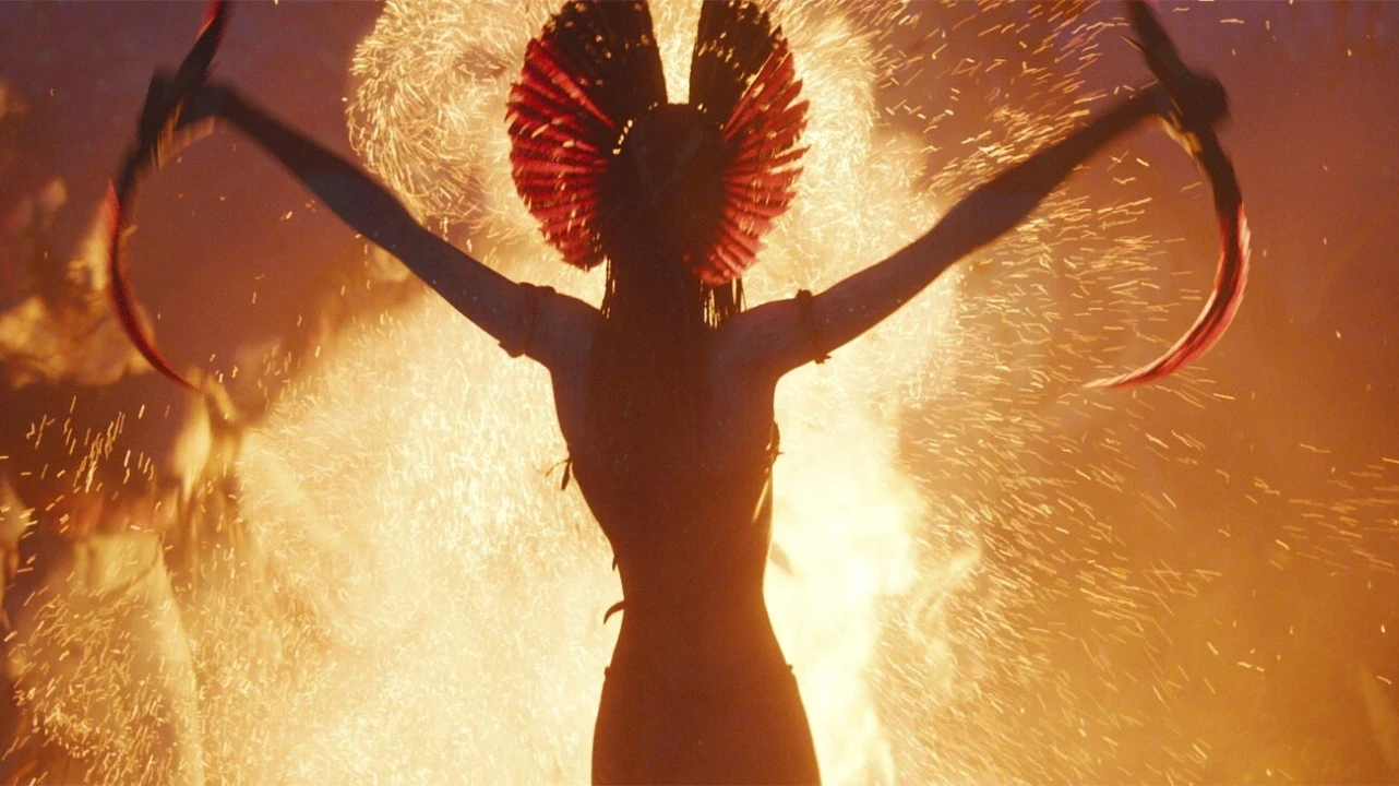 A silhouetted figure with outstretched arms stands against a vibrant, fiery background, holding curved, glowing red blades and wearing a headdress made of red feathers, evoking a sense of powerful energy and mystical drama.