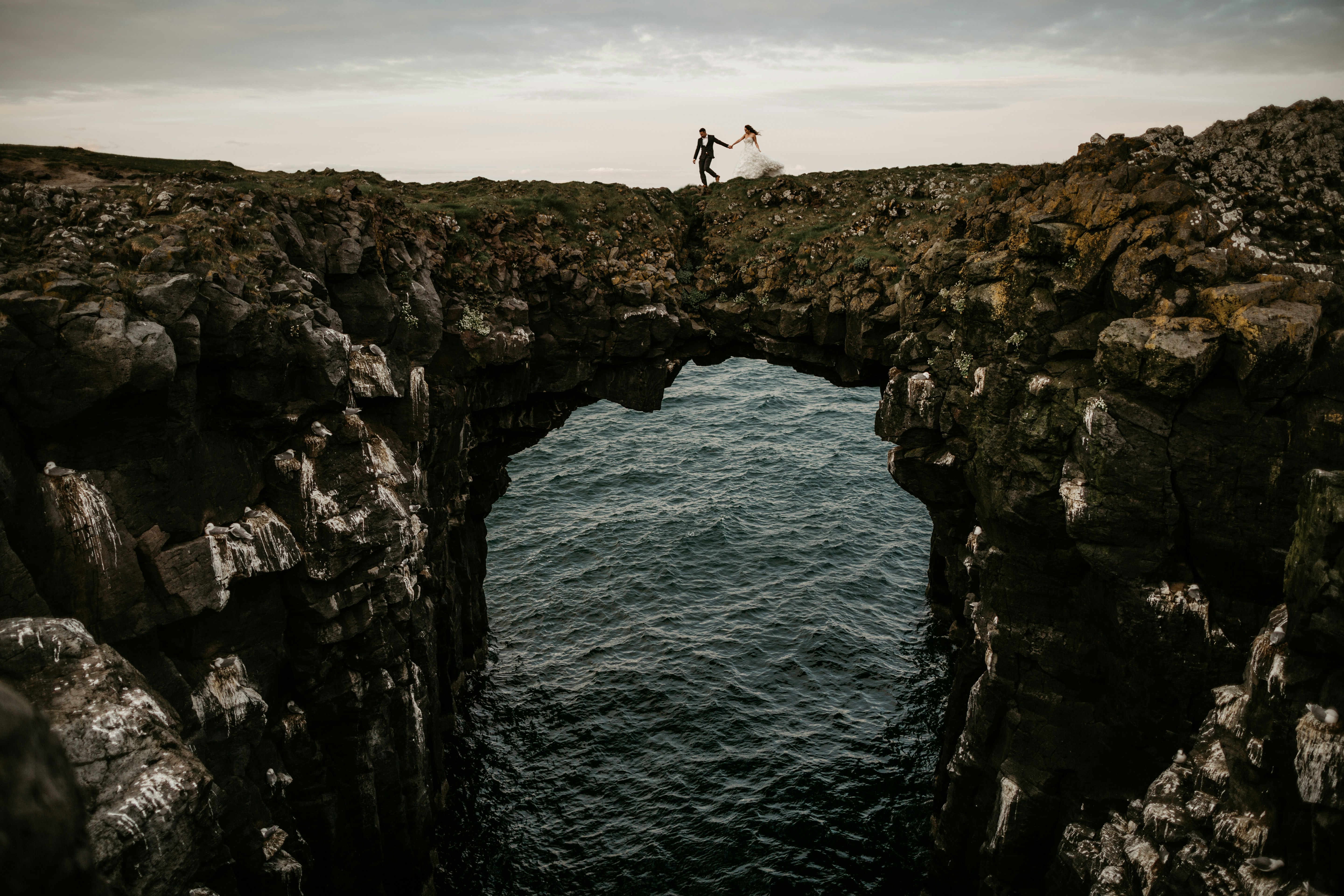 Wedding couple walking hand in hand above a stone arch in Iceland.