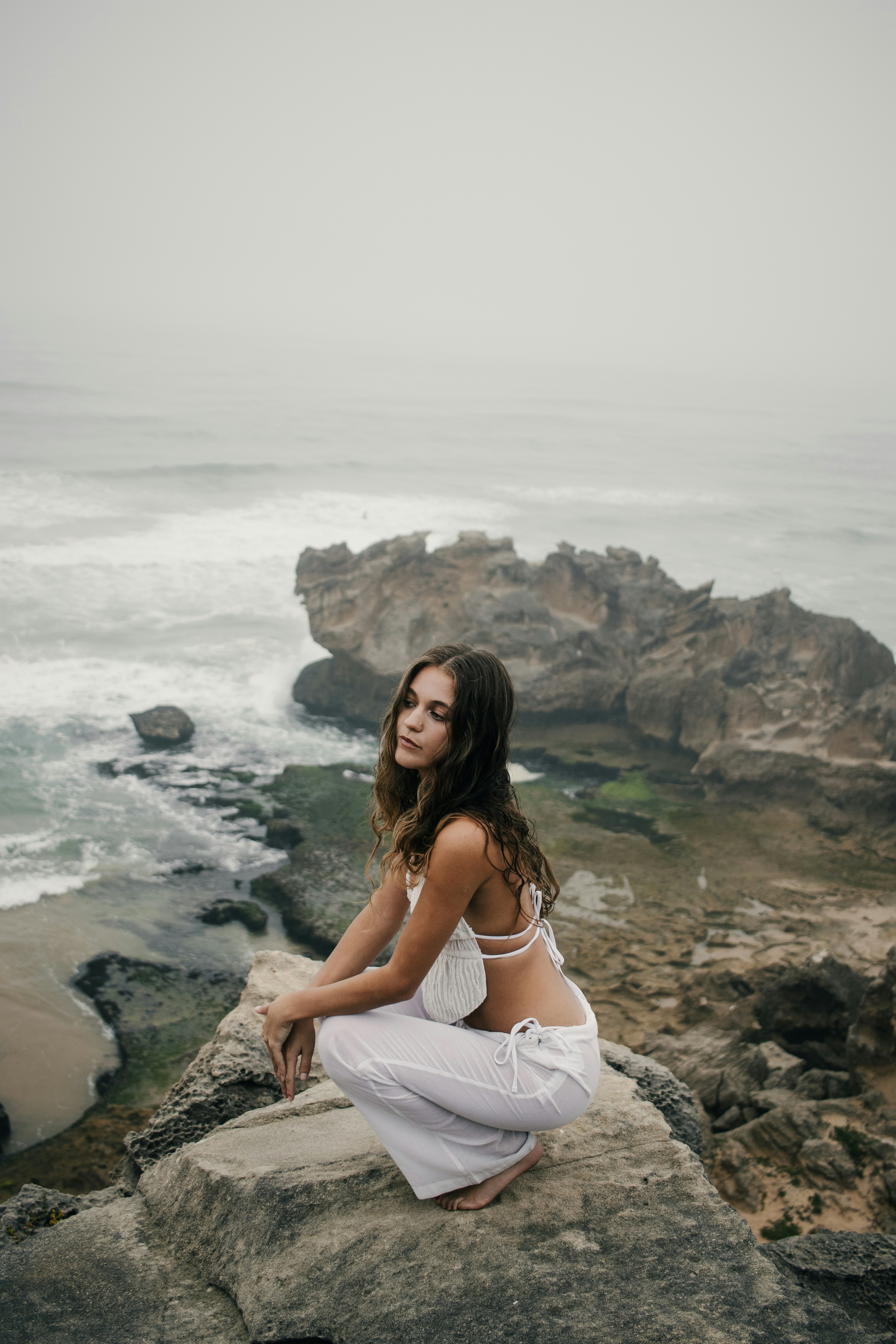 Woman in white clothes crouching on rocks by the ocean