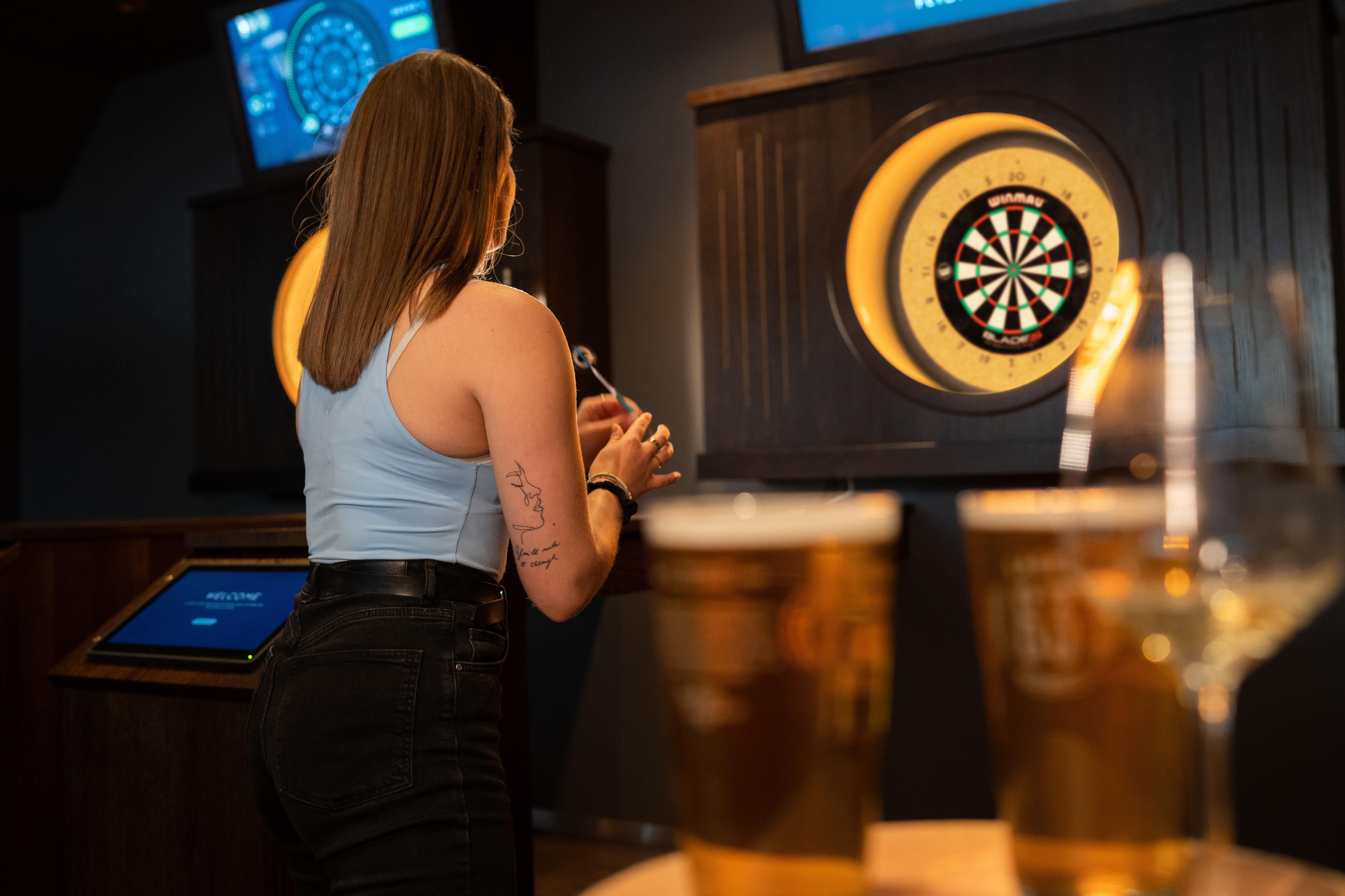 Una persona joven de cabello rizado sonríe a la cámara mientras está sentada frente a una computadora portátil sobre un fondo oscuro.