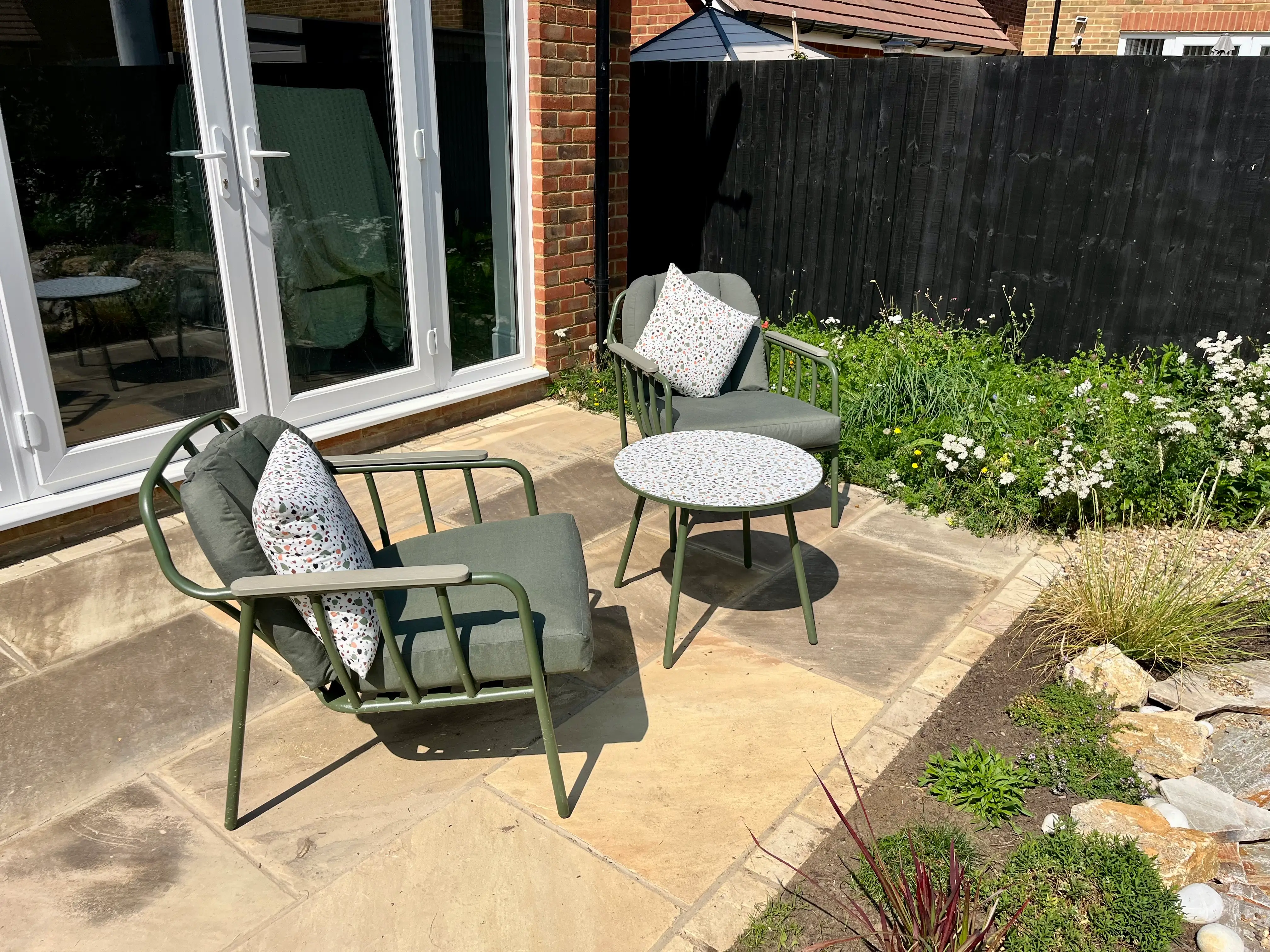Two outdoor chairs and a small table on a patio, surrounded by greenery and a sliding glass door in the background.