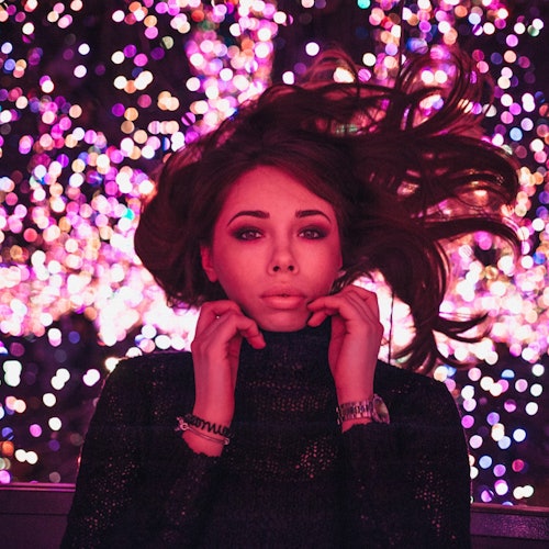 一位头发微乱的女性站在色彩斑斓的散景灯前摆姿势,双手放在衣领上。