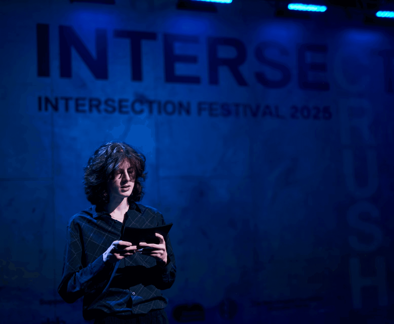 A young person stands alone onstage reading from a small booklet under moody blue lighting. Behind them, a large projection reads “INTERSECTION FESTIVAL 2025,” indicating a performance or presentation at the festival.