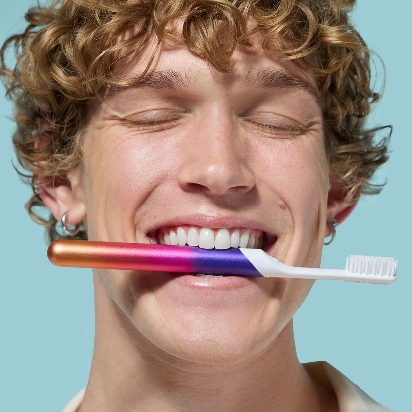 Smiling man with curly hair holding a toothbrush with an orange to purple gradient handle between his teeth