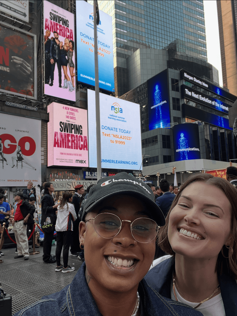 Ashleigh Warren and her partner Jessica smile for a selfie in Times Square, with bright digital billboards in the background featuring their HBO Max Show, Swiping America.