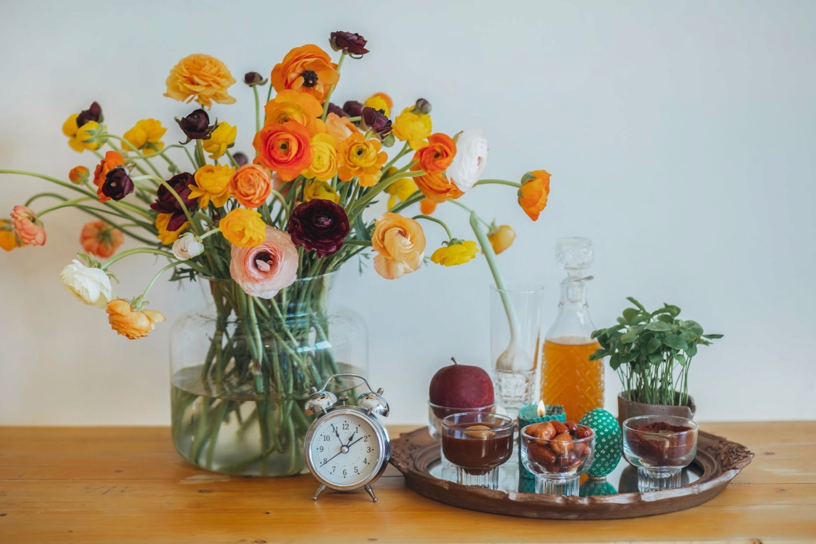 A vase of orange and yellow flowers and an alarm clock on a table, next to a tray containing the seven elements of the traditional Haft-Sin table (for Nowruz, Persian New Year)