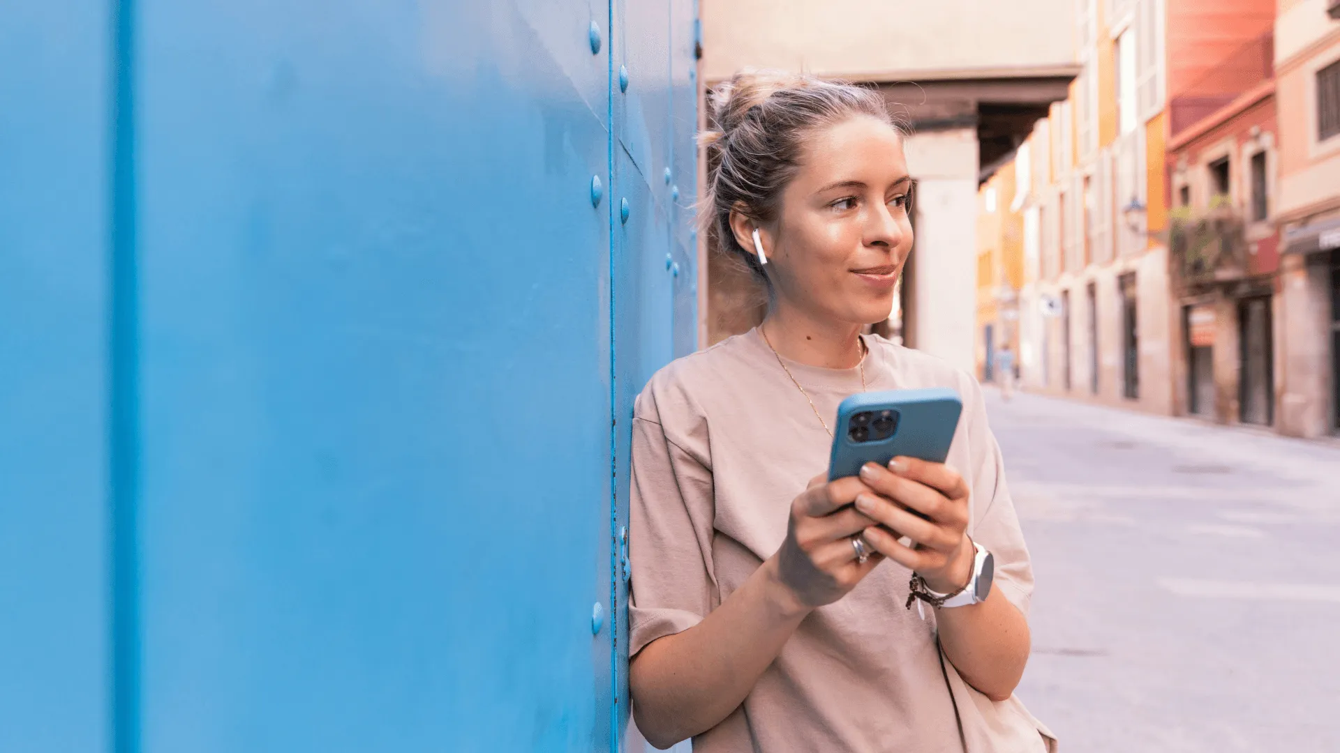 A woman with earbus in leans agains a blue wall, a phone in hand