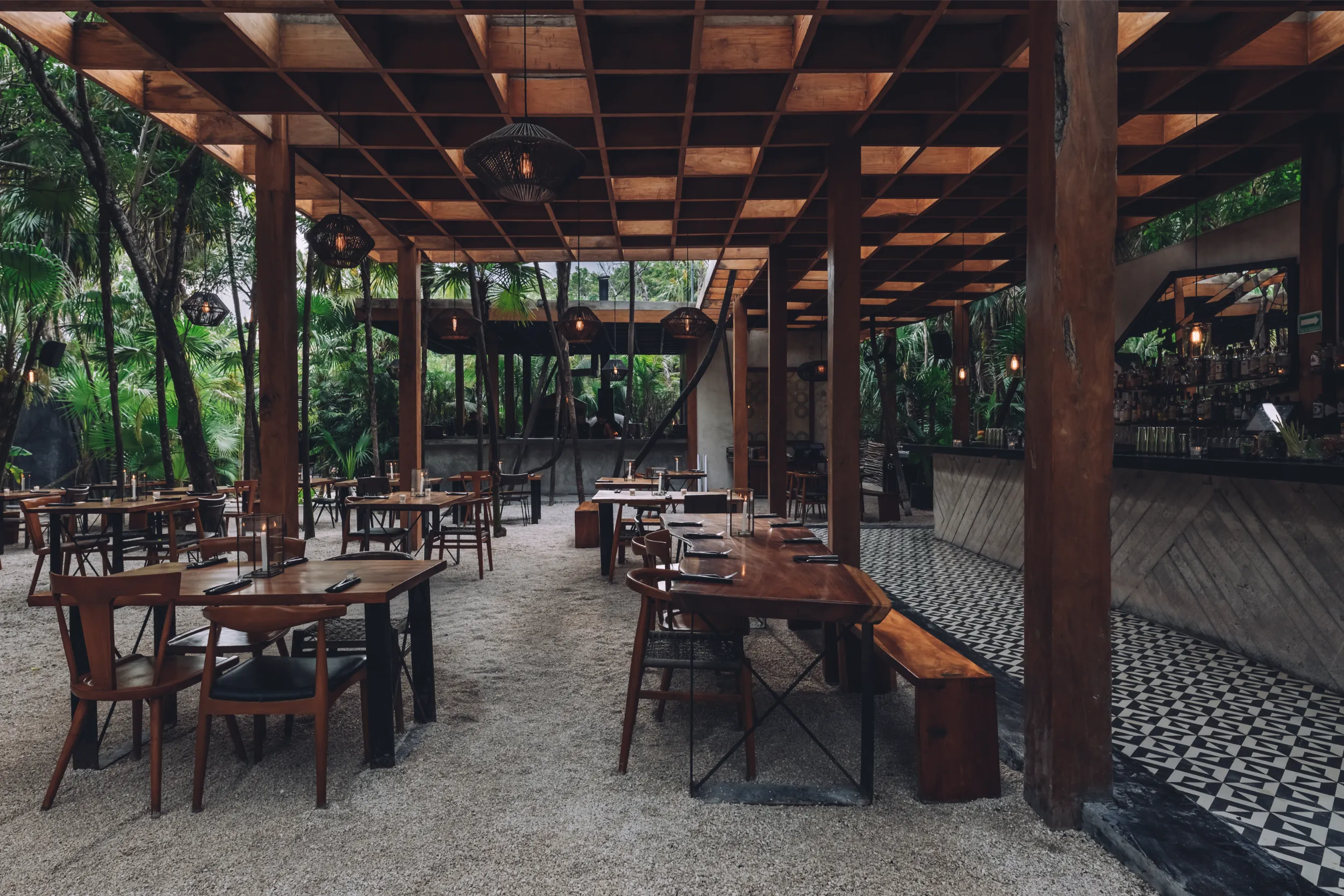 Interior atmosphere of the restaurant featuring native wooden furniture, gravel floors, and warm filament lighting under the wooden canopy.