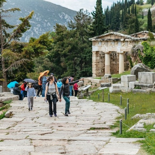 Pessoas caminhando por um caminho de pedra com ruínas antigas, árvores e montanhas ao fundo.