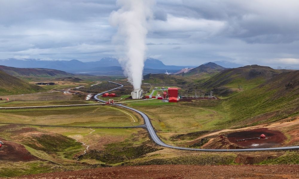 View of the Krafla geothermal power station in Iceland, with steam rising from geothermal facilities surrounded by green volcanic hills, winding roads, and distant mountains under cloudy skies.