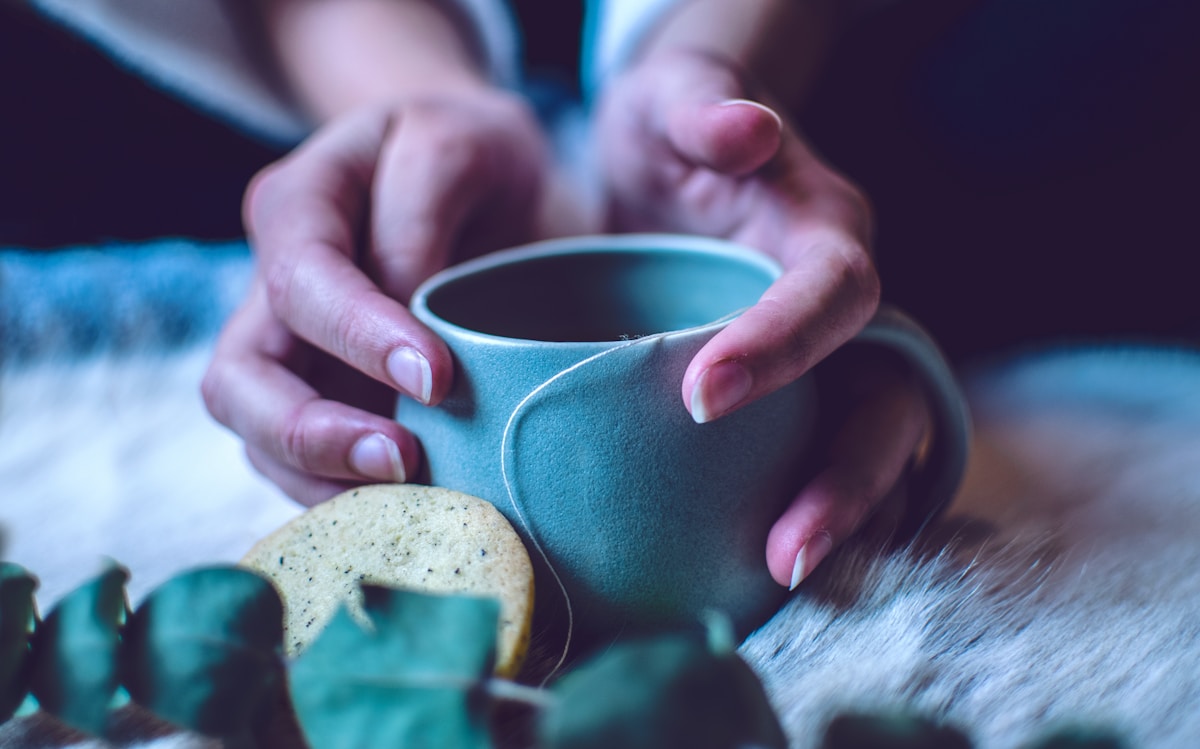 Hands gently holding a small teal mug beside a biscuit, evoking the warm, comforting meals that ease early refeeding discomfort