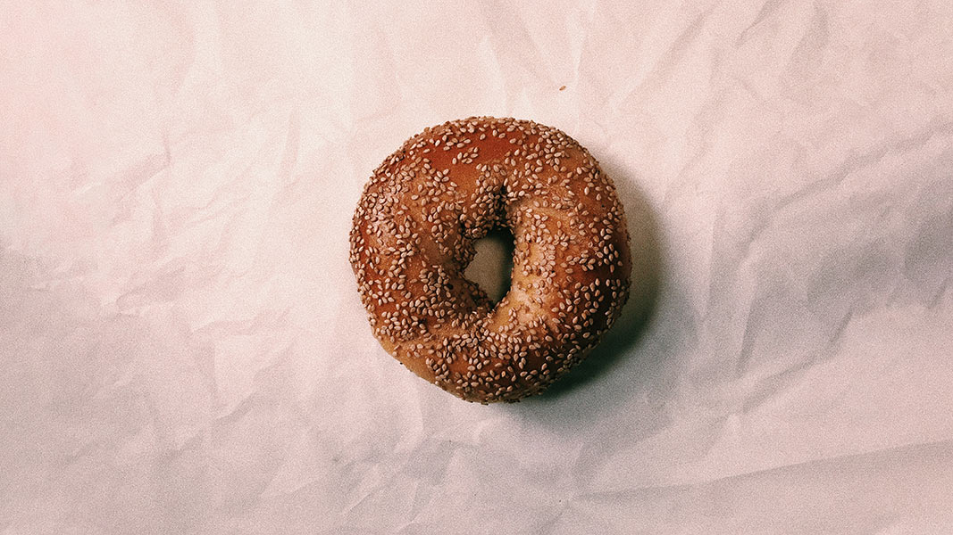 A single, light brown sesame seed bagel rests on a piece of off-white crumpled paper. The bagel is positioned centrally in the frame and has a hole in the center. The paper has a slightly textured appearance with visible wrinkles. No other objects or text are present in the image.