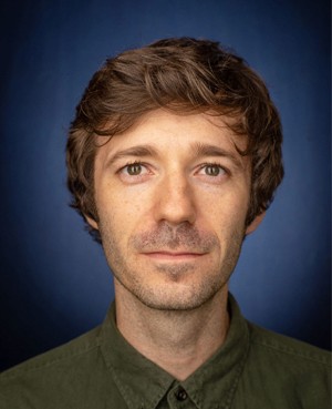 A young man with short, curly hair wearing a green shirt, set against a dark blue background.