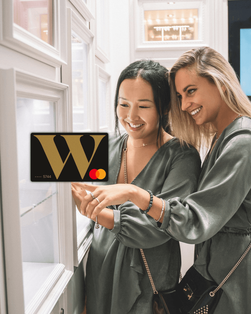 Two smiling friends shopping together with a black and gold Wealthsimple Cash Mastercard graphic overlaid, representing the ultimate zero-fee spending and bill-splitting card for newcomers in Canada.