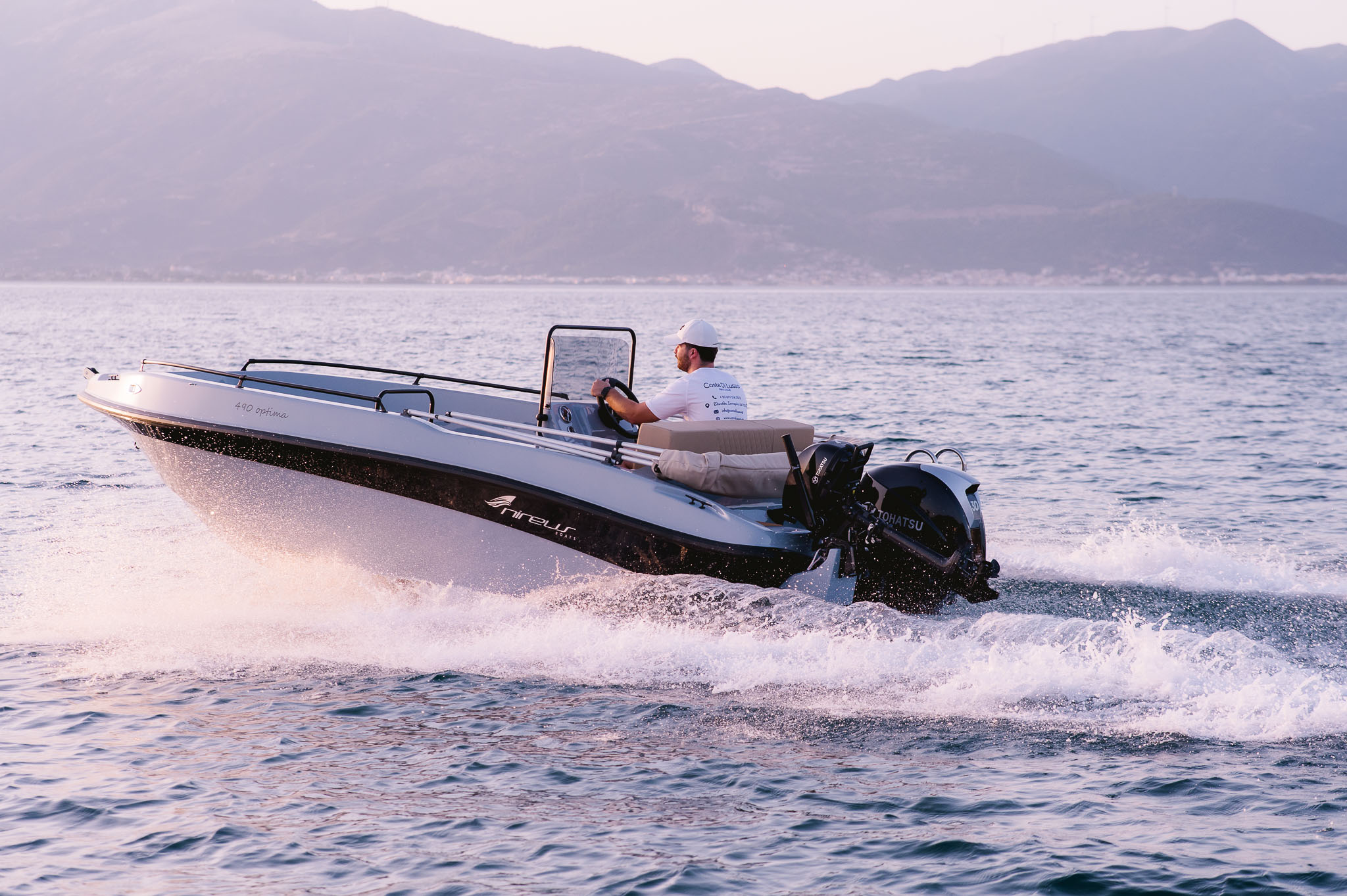 Nireus Optima Aura speedboat cruising at high speed with captain at helm, creating wake spray against Cycladic mountain backdrop.