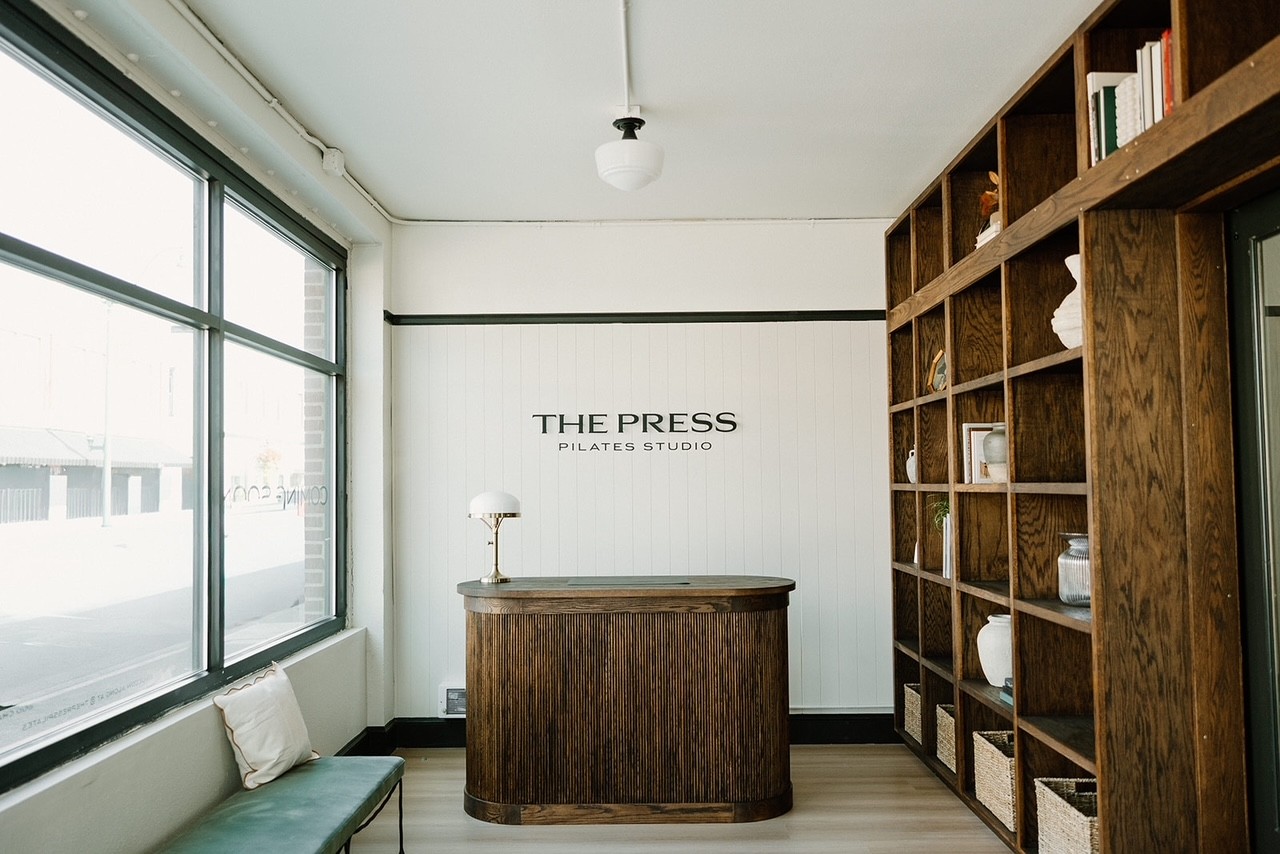 Reception area at The Press Pilates featuring a custom wall-mounted logo sign, curved wood desk, and warm, heritage-inspired interior details