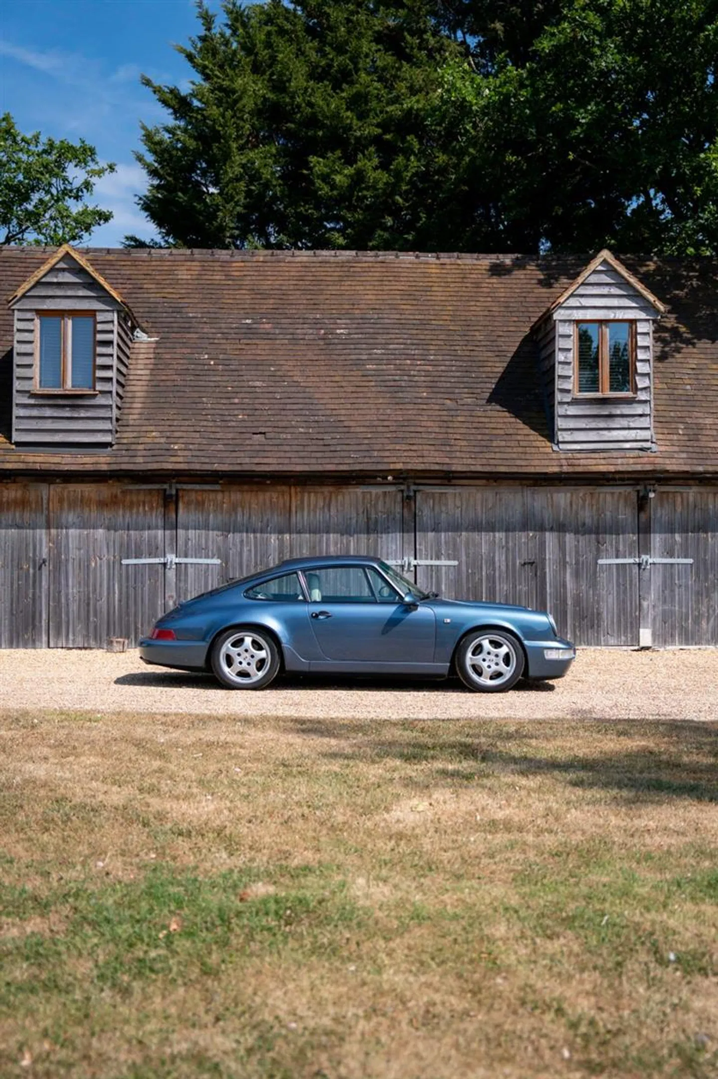 Side profile of the Baltic Blue 964 Carrera 2 in front of a wooden barn