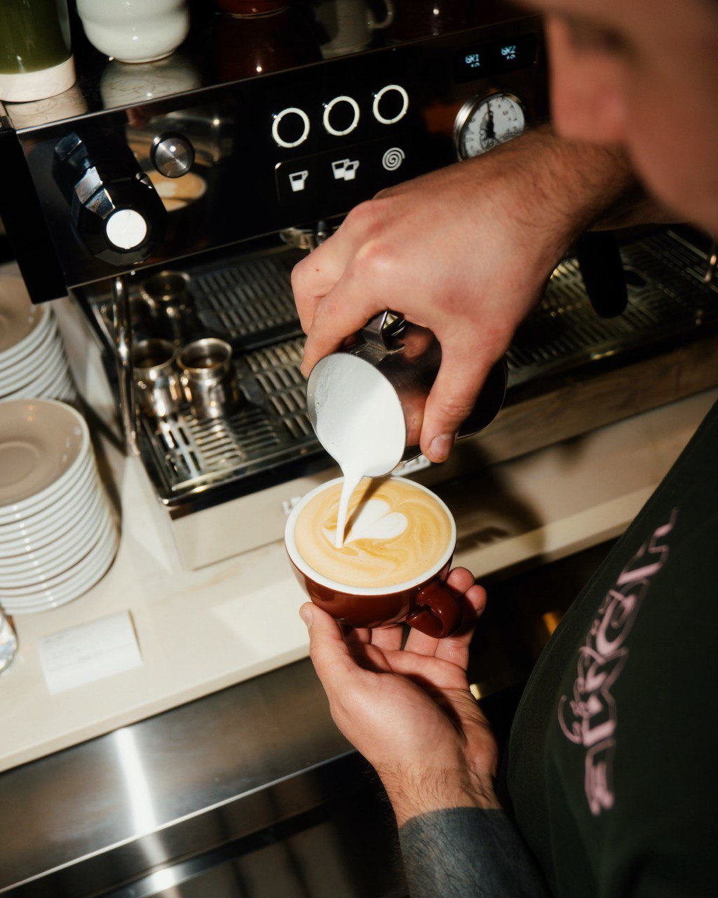 A barista skillfully pours steamed milk into a cup of espresso, creating an intricate latte art heart design on top of the rich, creamy coffee, while standing next to a shiny espresso machine.