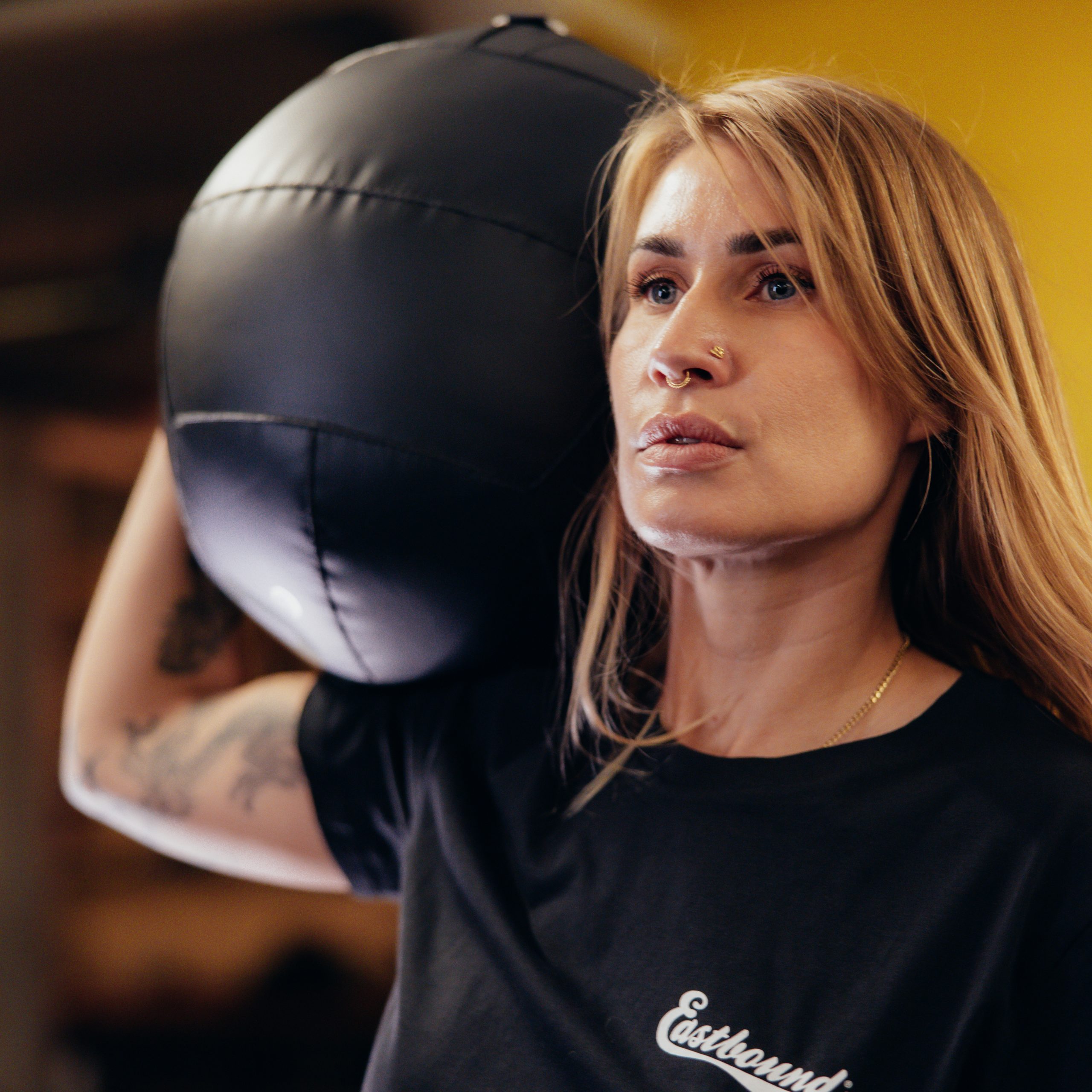 A focused woman holds a medicine ball overhead, training in a gym setting with a determined expression.