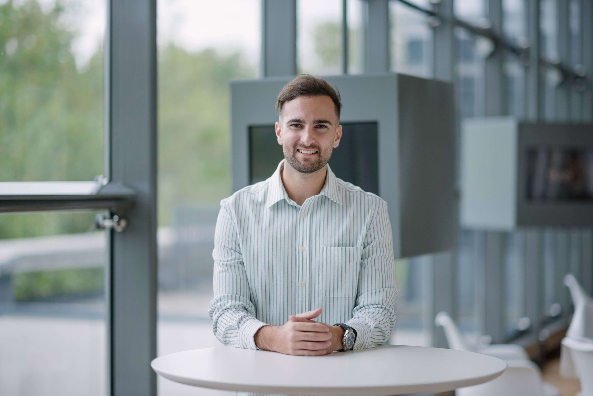 Young man from Team standing confidently at a table in a bright, modern office setting.