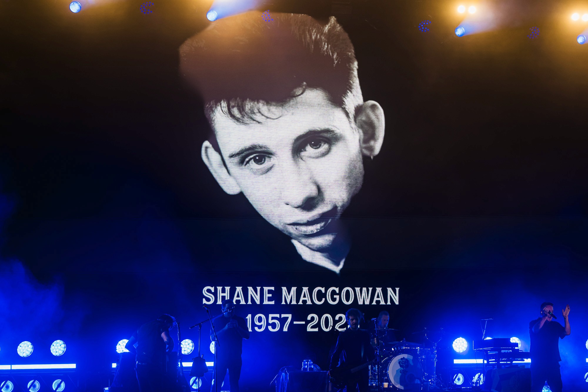 Hommage scénique à Shane MacGowan, projeté derrière les musiciens sous des lumières bleues.