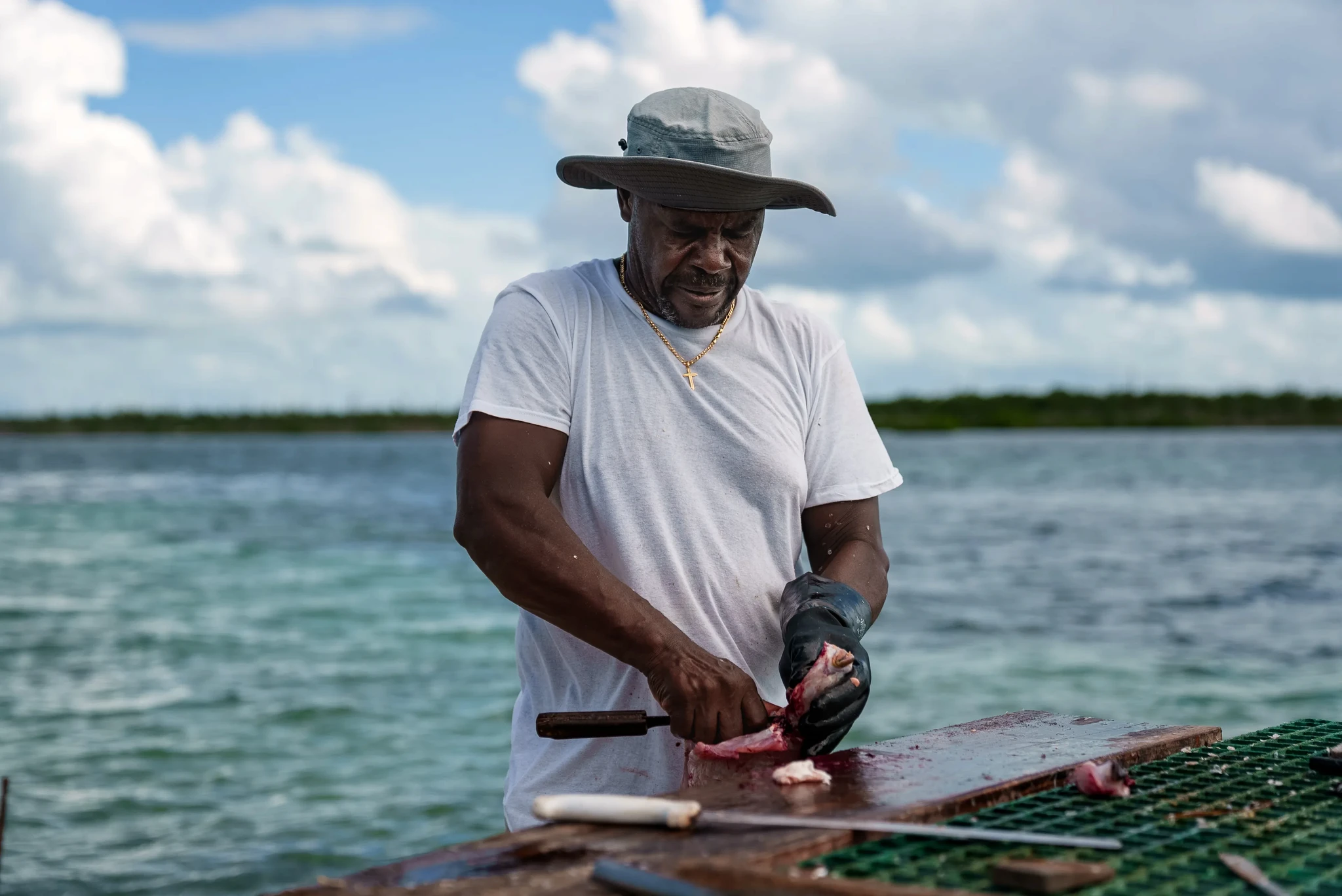 Man in a wide-brimmed hat and white t-shirt filleting a fish on a wooden surface outdoors, with a calm body of water and cloudy sky in the background. He is focused on his work, wearing a black glove on one hand and a gold cross necklace around his neck.