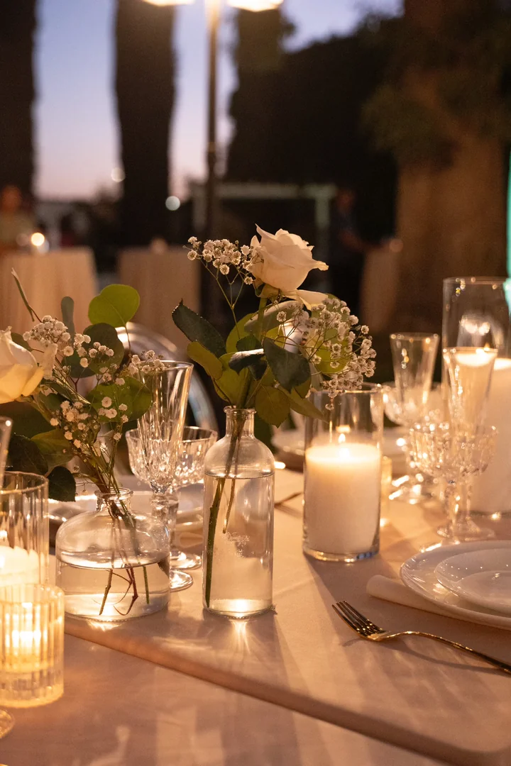 Long dinner table at dusk with lit white candles, crystal glasses, and bud vases of white roses and small white blooms