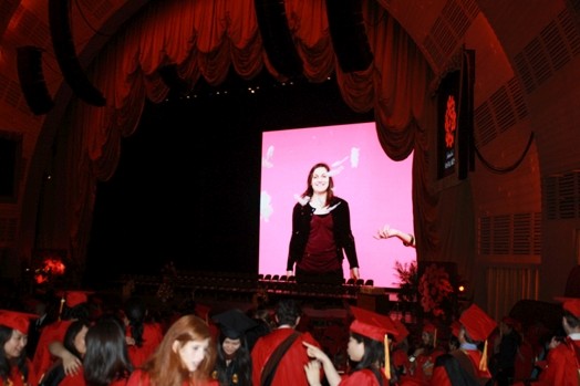 A photo of a theatre stage on which a picture of a girl is projected, while some students talk in the audience.