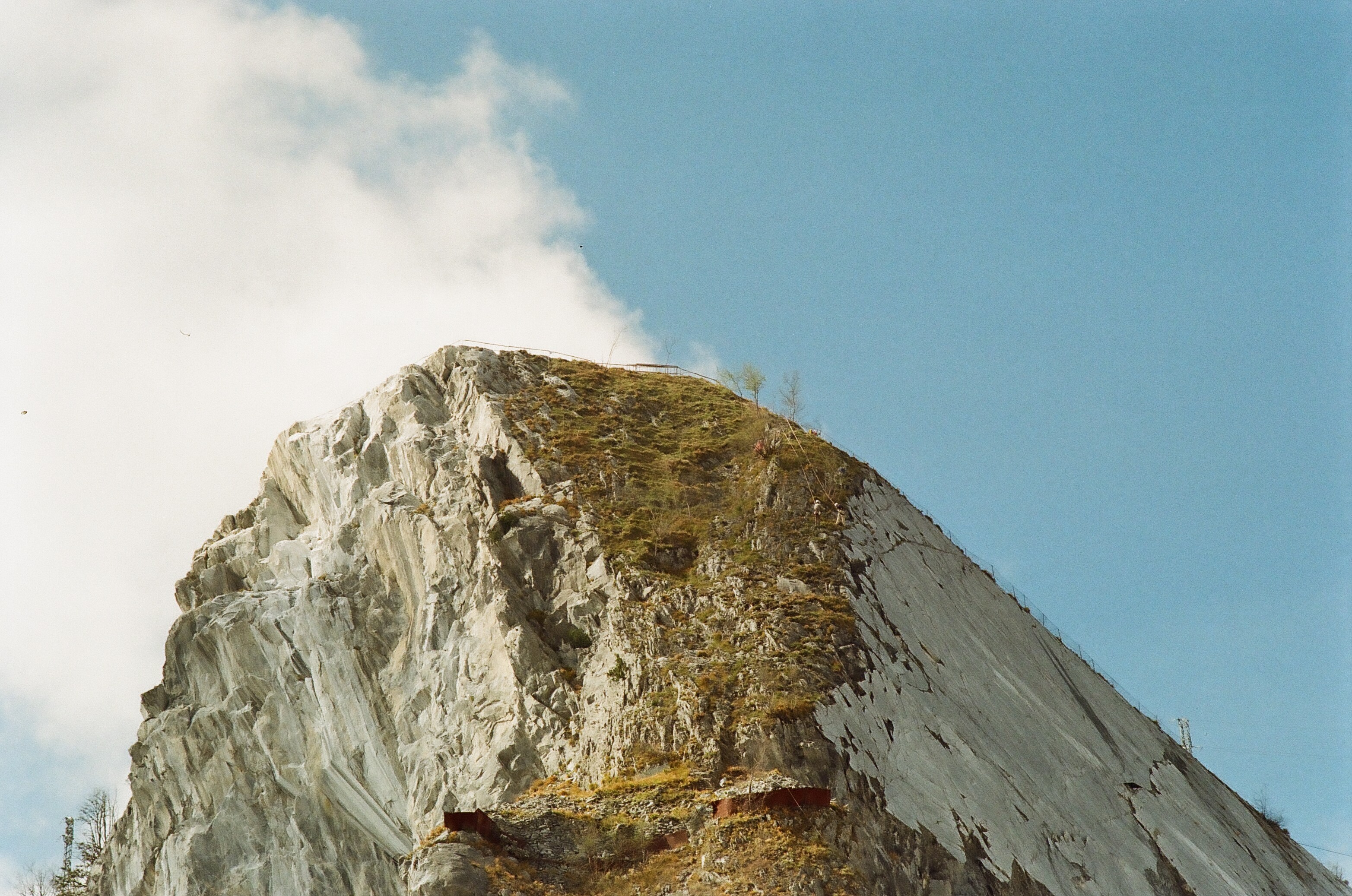 Sommet d'une montagne de marbre sur laquelle on aperçoit en tout petit des hommes encordés travaillant dans une pente escarpée