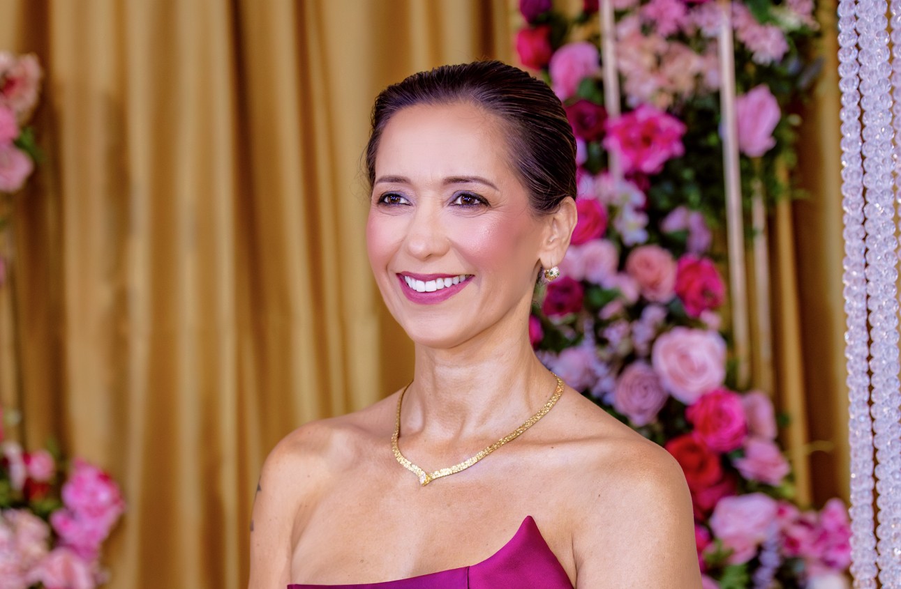 Foto da Maria Moreno, uma mulher elegante com um lindo sorriso. Ao fundo vamos uma parte do espaço de um evento, cortinas douradas e decoração floral com vários tons de rosa.