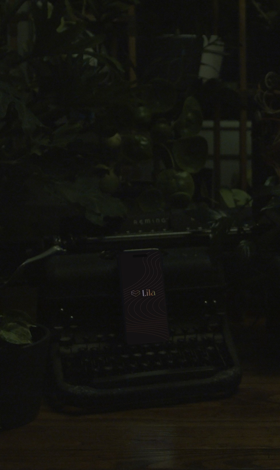 A photo of a dark office with a laptop and a mobile phone set atop a typwriter and surrounded by plants