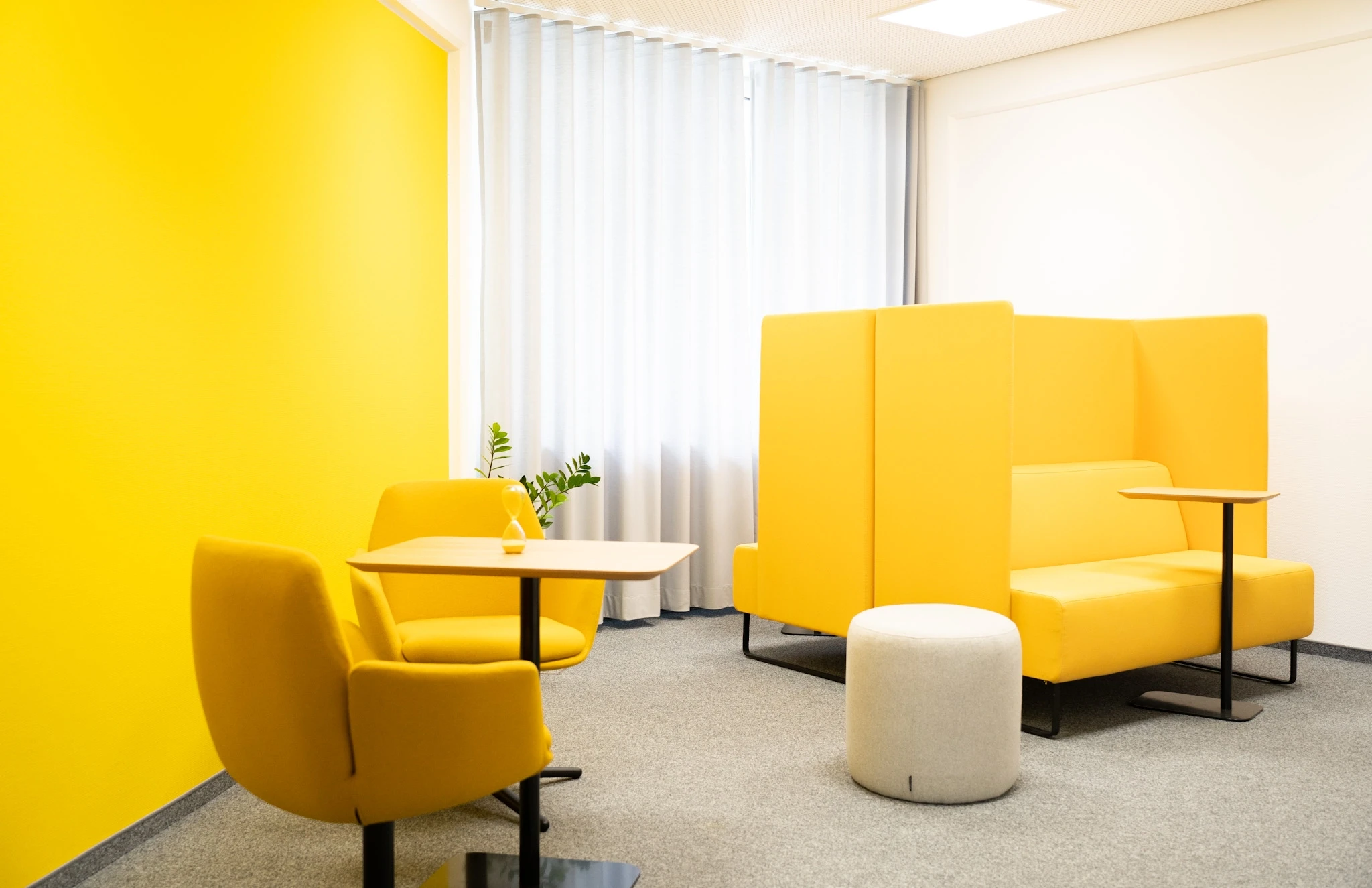 Yellow lounge room at NFON with a bright yellow wall, two yellow armchairs around a small wooden table, a yellow high-back sofa booth with privacy screens and a round white pouf.