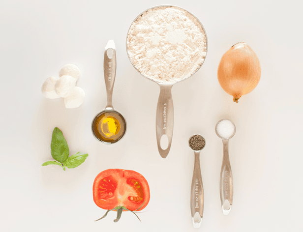 The image shows various ingredients arranged on a white background. On the left side, there are three white balls, a green basil leaf, and a halved ripe red tomato. In the center, there are two measuring spoons: one containing an amber liquid (likely oil) and the other filled with white flour. To the right of the flour, a whole onion, a black peppercorn, and a small pile of salt are visible. Two measuring spoons are also present: one measuring 1/2 teaspoon of salt and the other measuring 1/4 teaspoon of pepper.