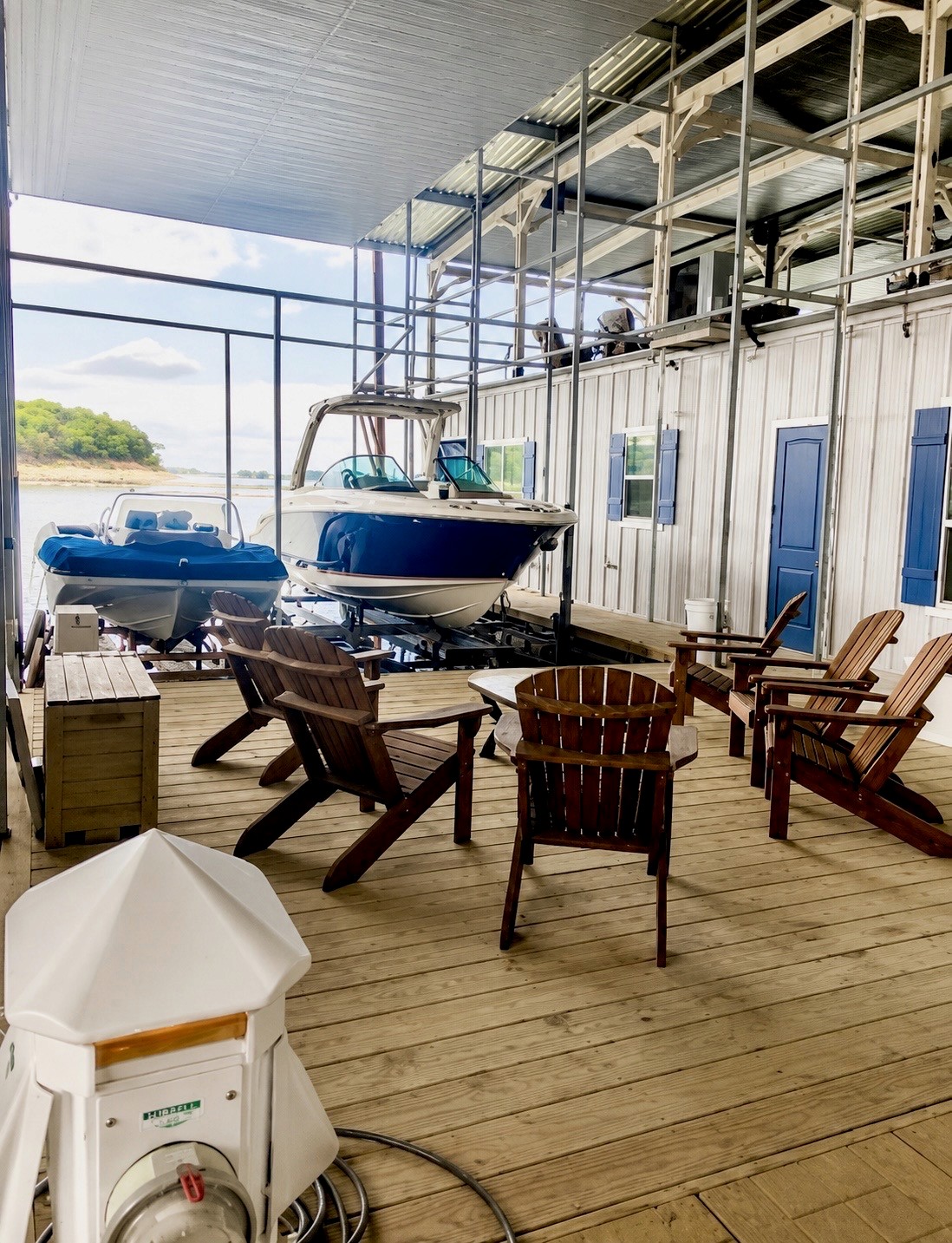 A cozy lakeside boathouse features a wooden deck with several Adirondack chairs arranged around a small table, overlooking two docked boats under a metal-roofed shelter with a scenic view of calm waters and a distant tree-lined shore.