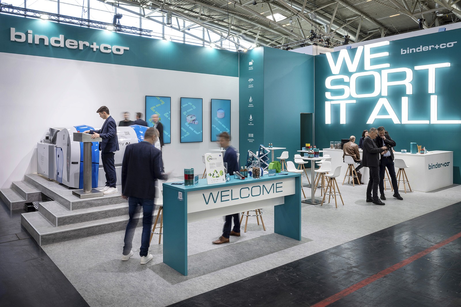 Der bauma 2025 Messestand von binder+co in ihrem Petrolblau mit dem großen Slogan "We sort it all" im Hintergrund. Mehrere Personen stehen herum, links ist eine Clarity-Maschine zu sehen