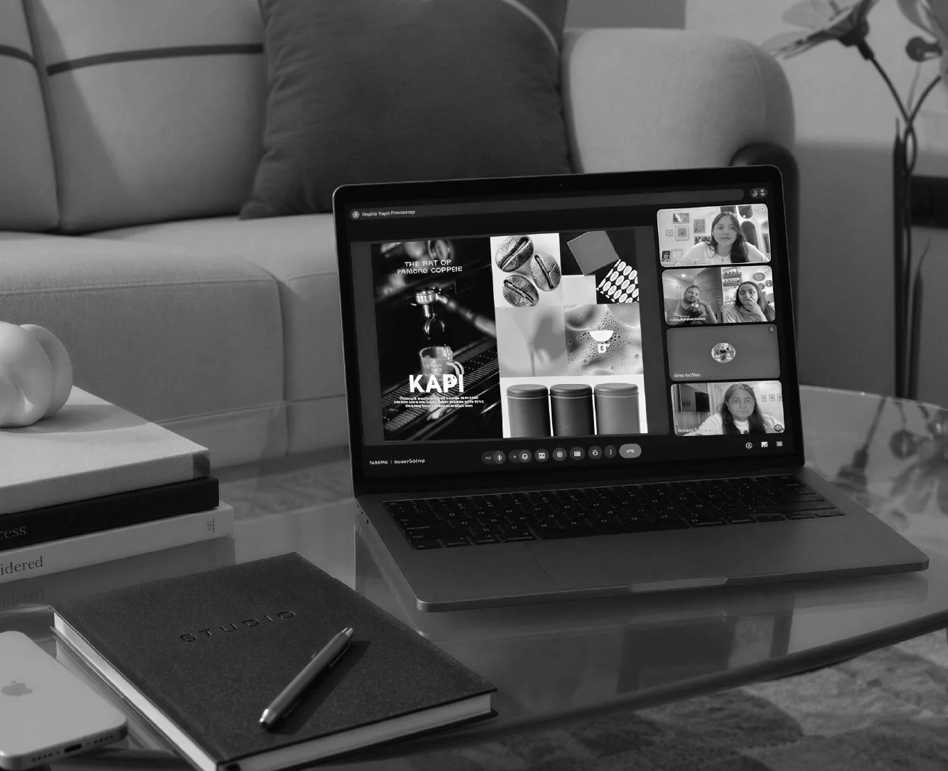 A laptop displaying a virtual meeting on its screen sits on a glass coffee table surrounded by a notebook, pen, and smartphone, with a sofa and decorative items in the background, illustrating a modern home office setup.