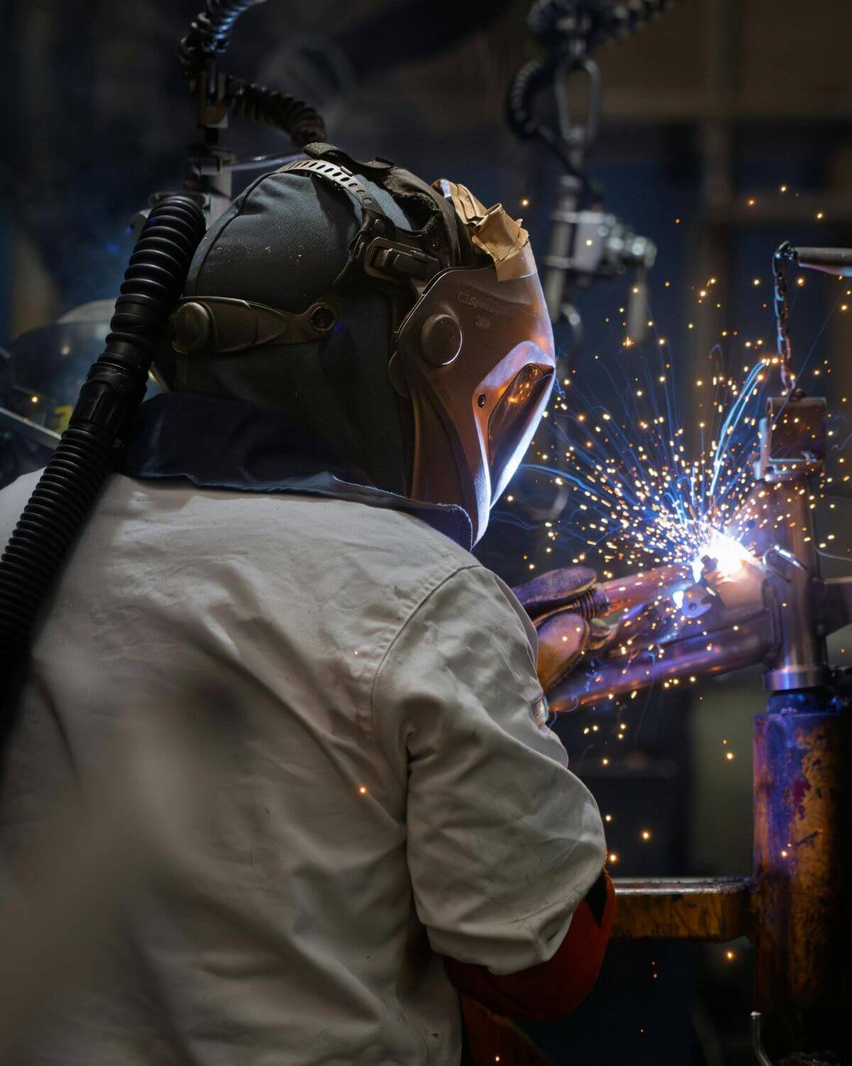 Welder in protective mask welding metal with bright sparks in steel fabrication workshop, demonstrating energy-intensive manufacturing operations that benefit from battery storage optimization