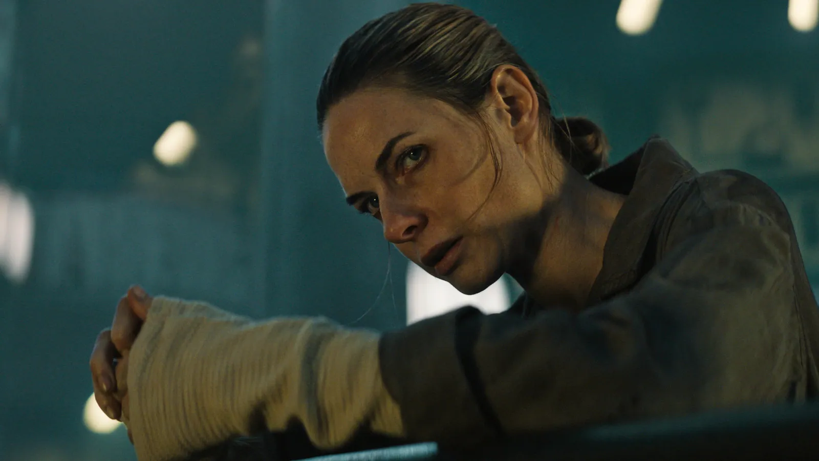 A woman with a determined expression, wearing a jacket, leans on a railing in a dimly lit industrial setting, her hands wrapped in white bandages.