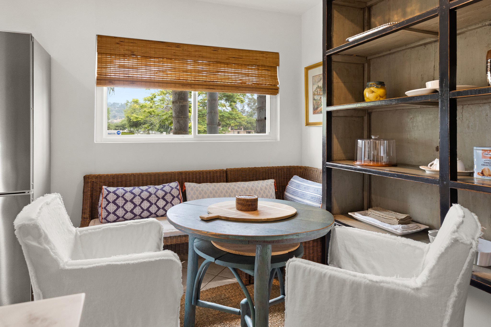 A bright breakfast nook with woven seating, soft layered cushions, and views of the palm-lined neighborhood.