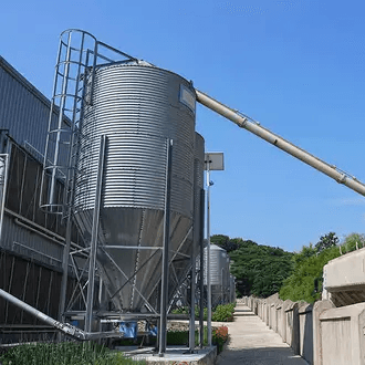 Large corrugated metal silos and an inclined conveyor belt system under a clear blue sky, illustrating efficient.