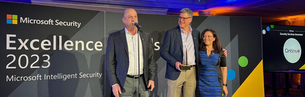 Drei Personen stehen bei einer Veranstaltung gemeinsam auf der Bühne, wobei eine Person in ein Mikrofon spricht. Sie halten eine Auszeichnung vor einem Hintergrund, auf dem 'Microsoft Security Excellence 2023' und das Logo von 'Onnrive' zu sehen sind.