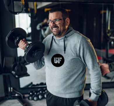 Een man traint enthousiast met dumbbells in de sportschool, gekleed in een 5 Day Fast hoodie.