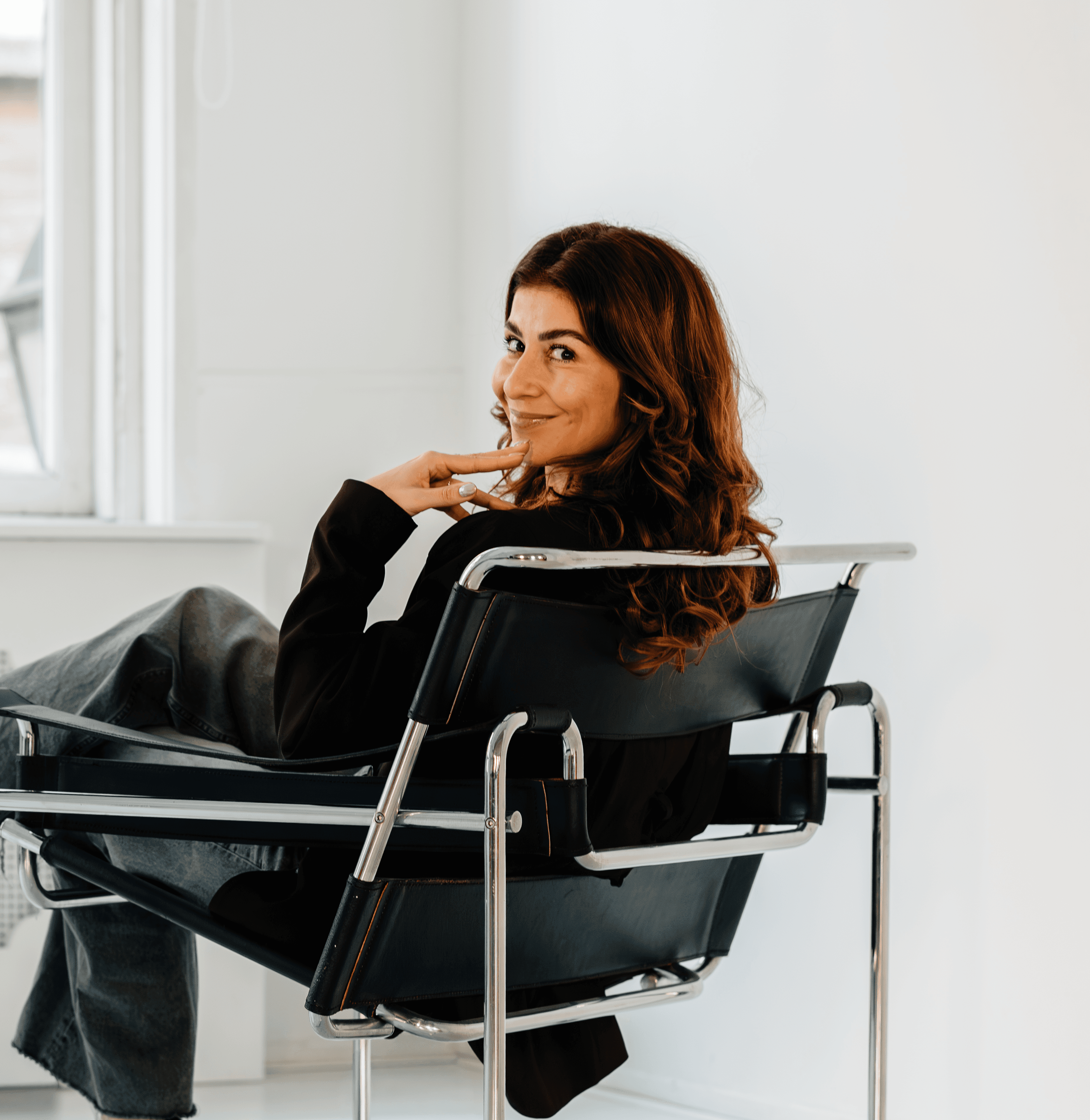 A woman with long brown hair sits casually in a modern chair, looking over her shoulder, in a bright room.